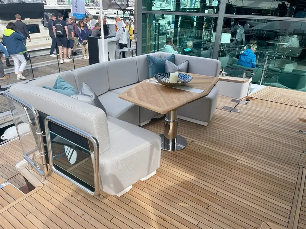 Stylish outdoor seating area on a boat, featuring a grey sofa, wooden table, and decorative items, with people nearby.